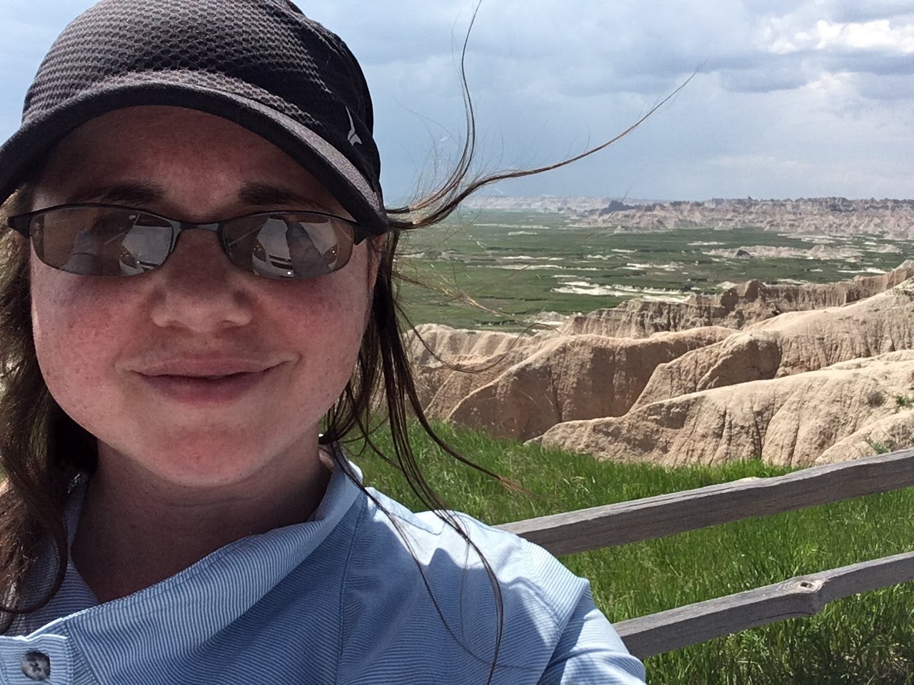 Ashley Book wearing sunglasses and a cap with a scenic view of cliffs and fields in the background
